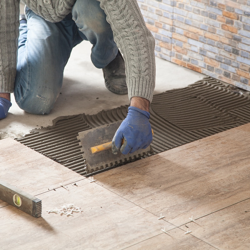 Artisan appliquant de la colle à carrelage avec une truelle dentelée sur le sol, préparant la pose de carreaux beiges.