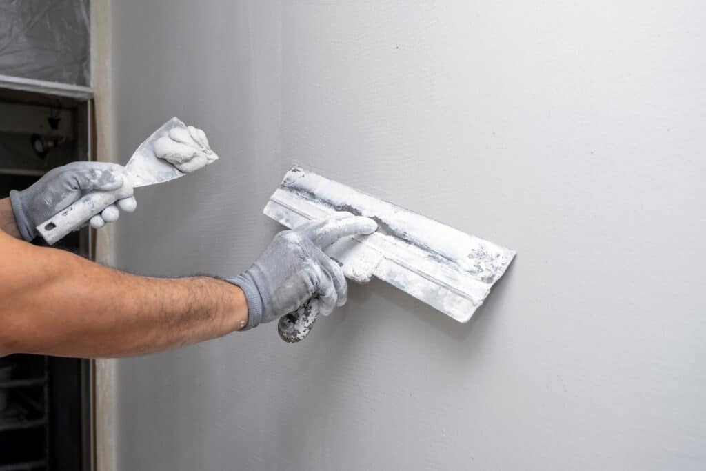 Application d'enduit de plâtre avec truelles : finitions murs Mains gantées appliquant de l'enduit blanc sur un mur gris clair avec deux truelles lors de travaux de rénovation.