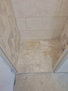 Intérieur de douche carrelé, murs beige texturé 3D et sol effet travertin beige/brun clair, cadre de porte gris au sol brut.