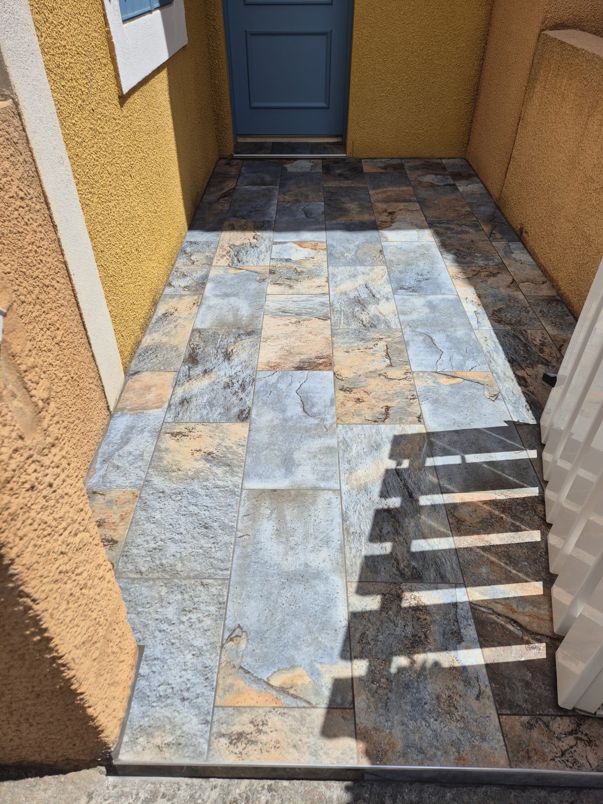 Entrée ensoleillée avec un sol carrelé imitation ardoise bleu/ocre et une porte bleu marine, ombres de barres horizontales projetées sur le carrelage.