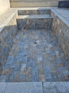 Piscine en construction carrelée avec des dalles imitation ardoise bleu-brun, évacuation centrale au sol et spots à carreler.