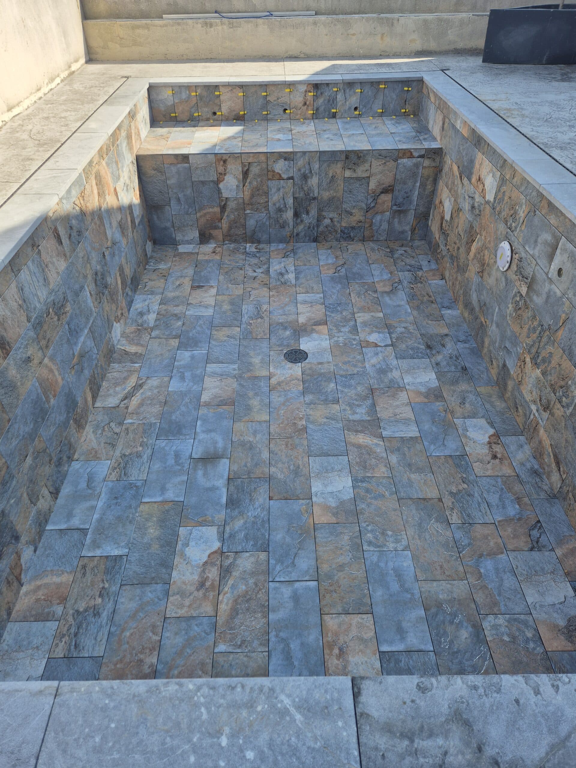 Piscine en construction carrelée avec des dalles imitation ardoise bleu-brun, évacuation centrale au sol et spots à carreler.
