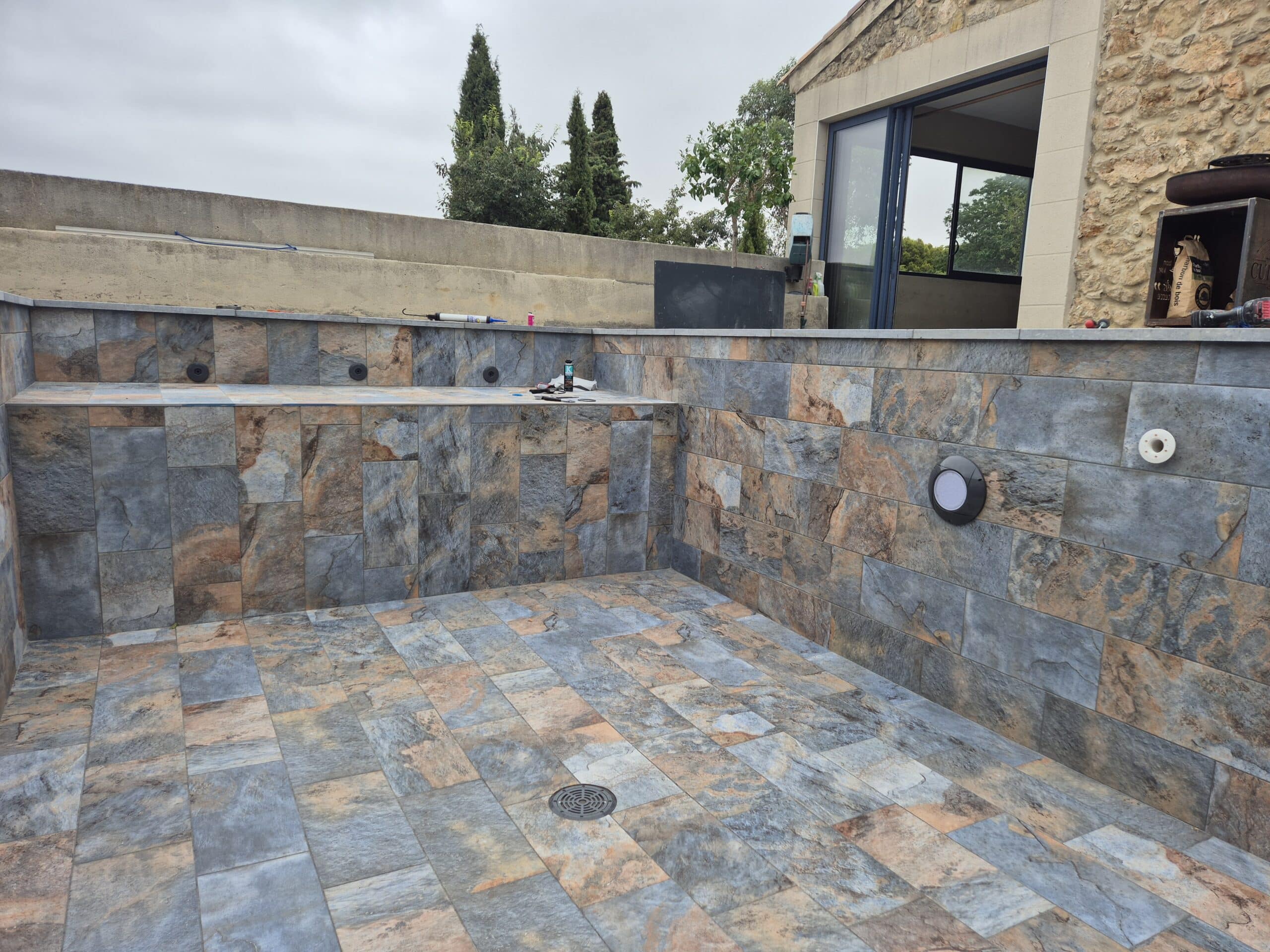 Piscine en construction, fond et murs recouverts de carrelage imitation ardoise bleu-brun. Un drain au sol est visible, près d'une maison en pierre.