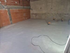 Chantier intérieur avec sol fraîchement coulé (chape ciment), murs en briques rouges alvéolées et murs en béton brut, présence de conduits gris et de tubes bouchonnés.