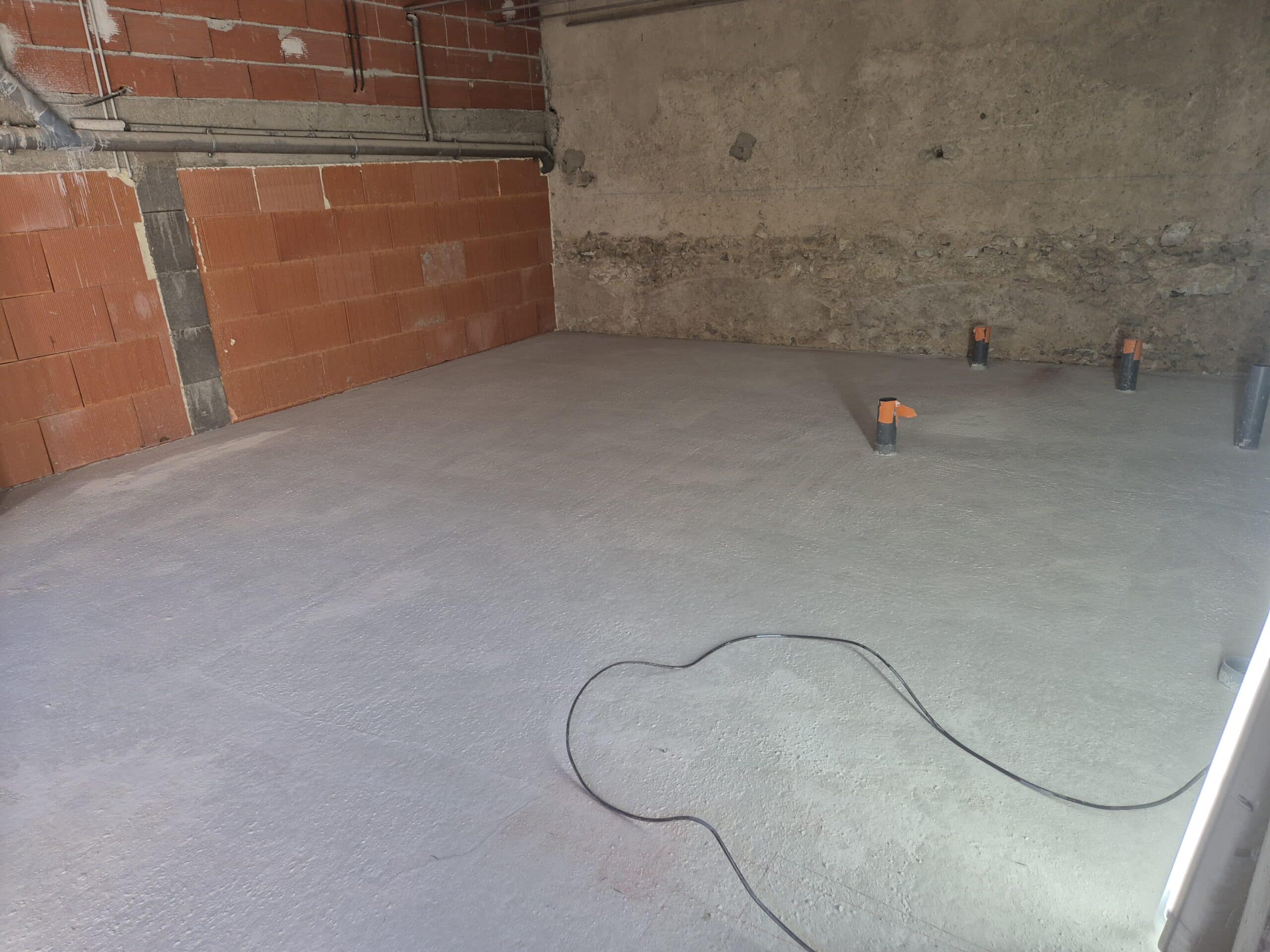 Chantier intérieur avec sol fraîchement coulé (chape ciment), murs en briques rouges alvéolées et murs en béton brut, présence de conduits gris et de tubes bouchonnés.