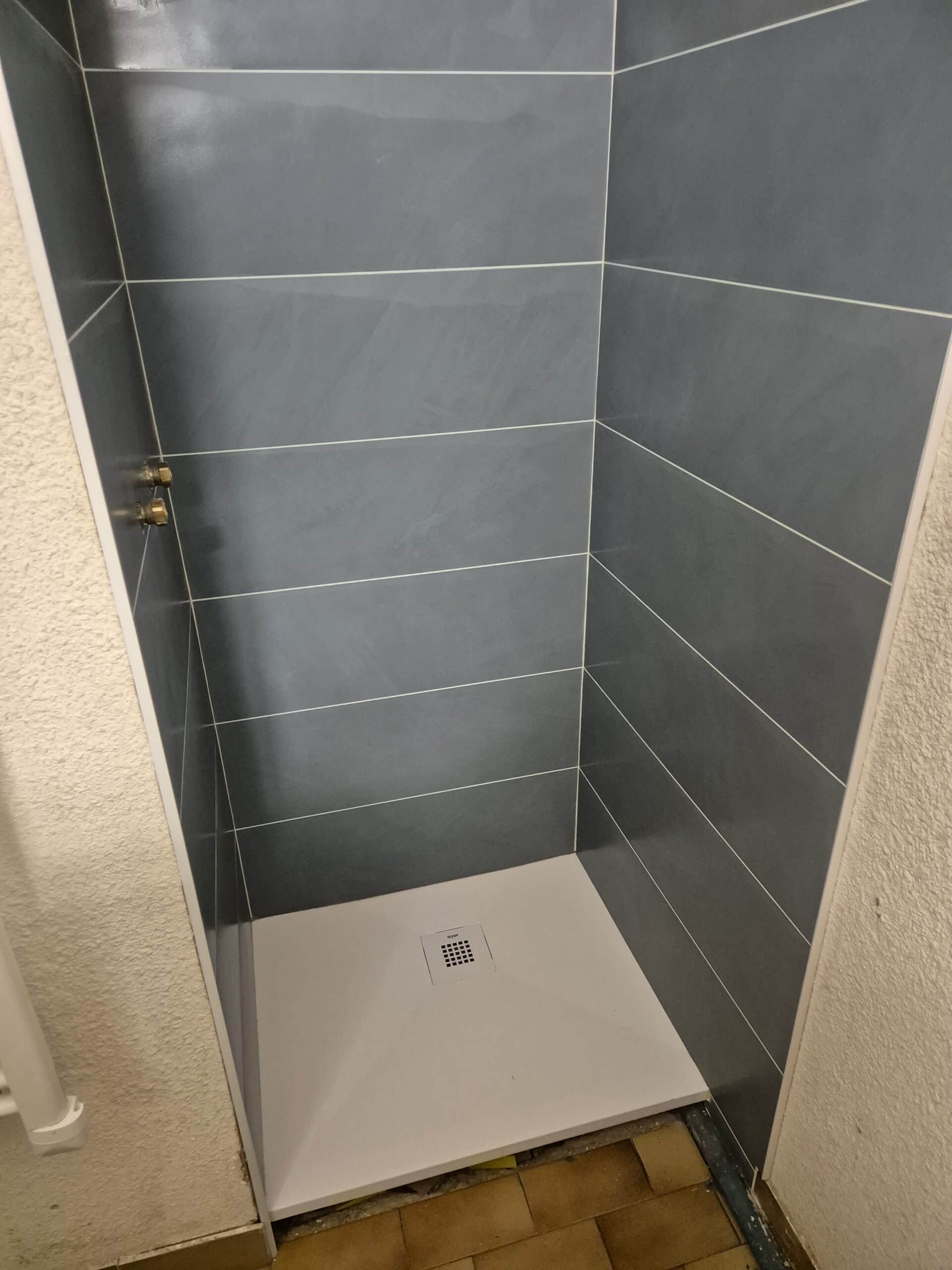 Douche moderne carrelée gris anthracite avec joints clairs et receveur blanc. Tuyauterie laiton visible à gauche. Drain portant la marque 'ayor'.