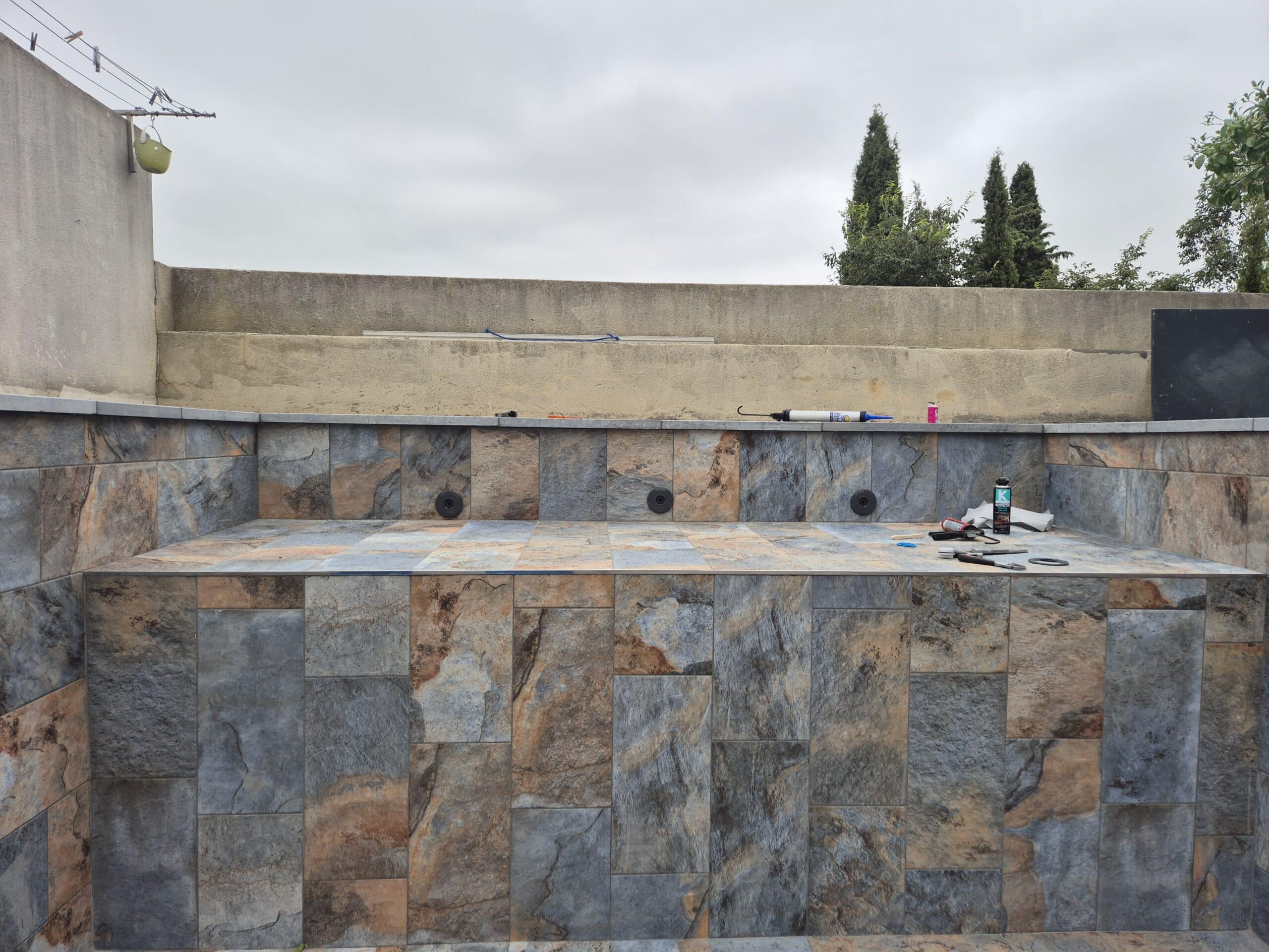 Fond de piscine vide carrelé avec des carreaux imitation ardoise multicolores (bleu, rouille) et des outils de calfeutrage posés sur la marche.