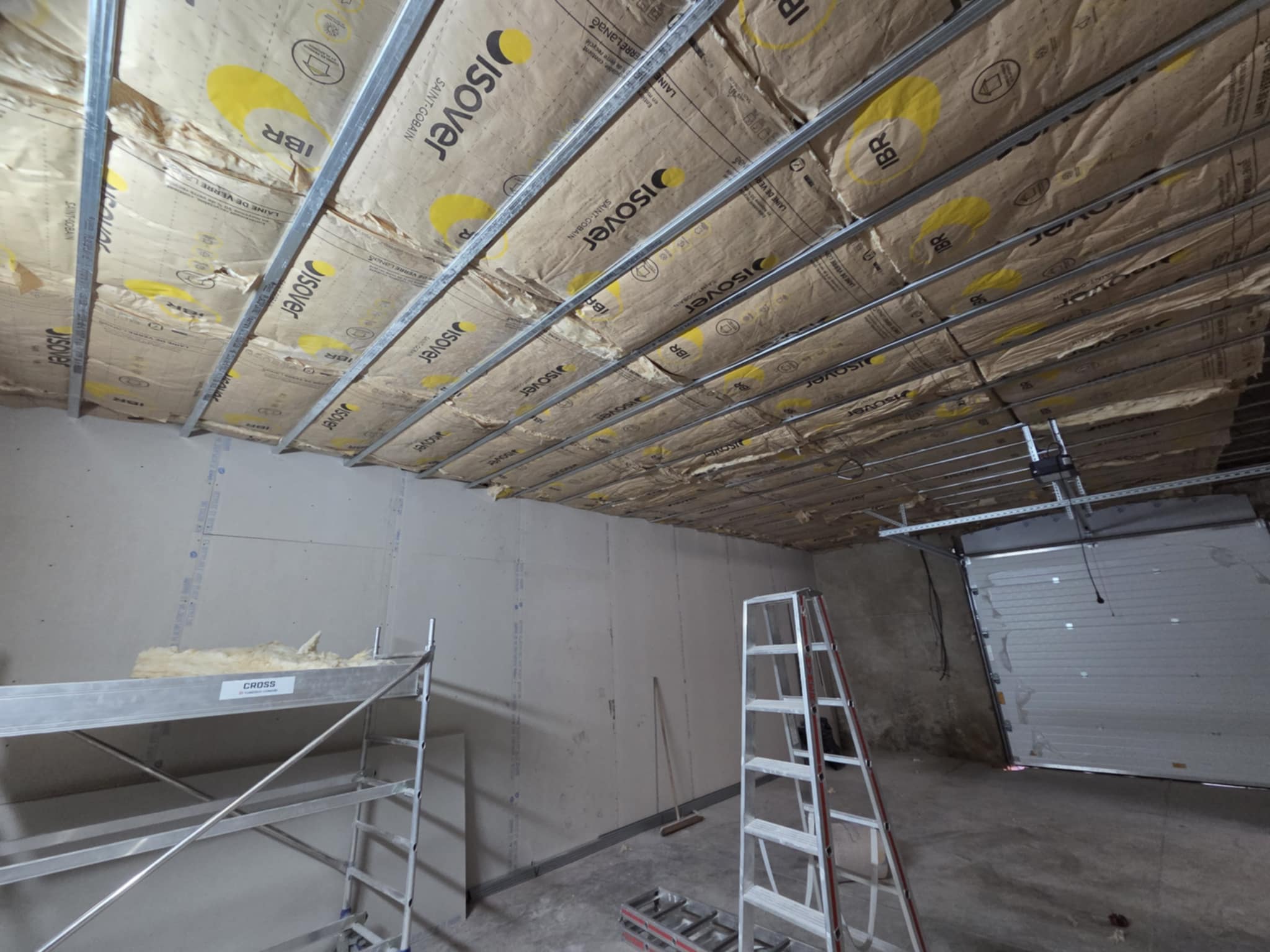 Isolation de plafond en laine de verre ISOVER entre ossatures métalliques. Chantier avec des plaques de placo et des échelles.