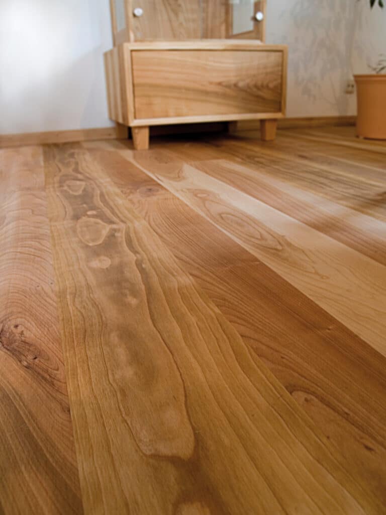 Plancher Bois Clair Chaleureux et Mobilier Assorti Plancher en bois clair et chaleureux, avec des veines prononcées, près d'une commode en bois assortie et d'un mur blanc avec une ombre de plante.
