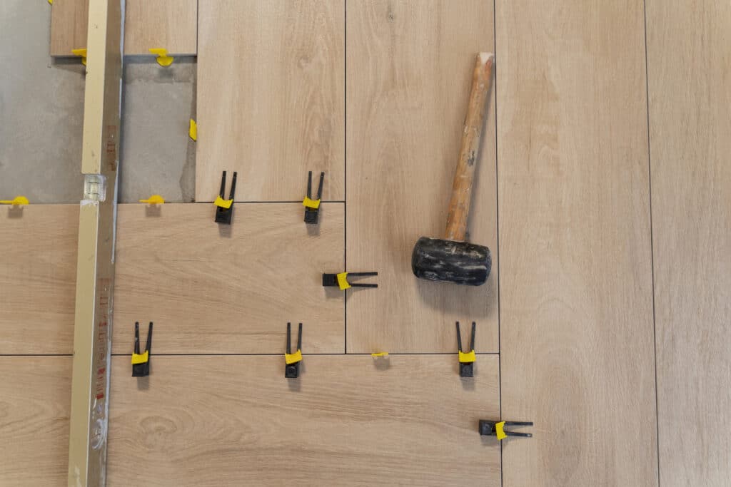 Pose Carrelage Imitation Bois : Outils de Nivellement Pose de carrelage imitation bois clair avec croisillons et niveleurs jaunes en cours, un maillet repose sur les carreaux.