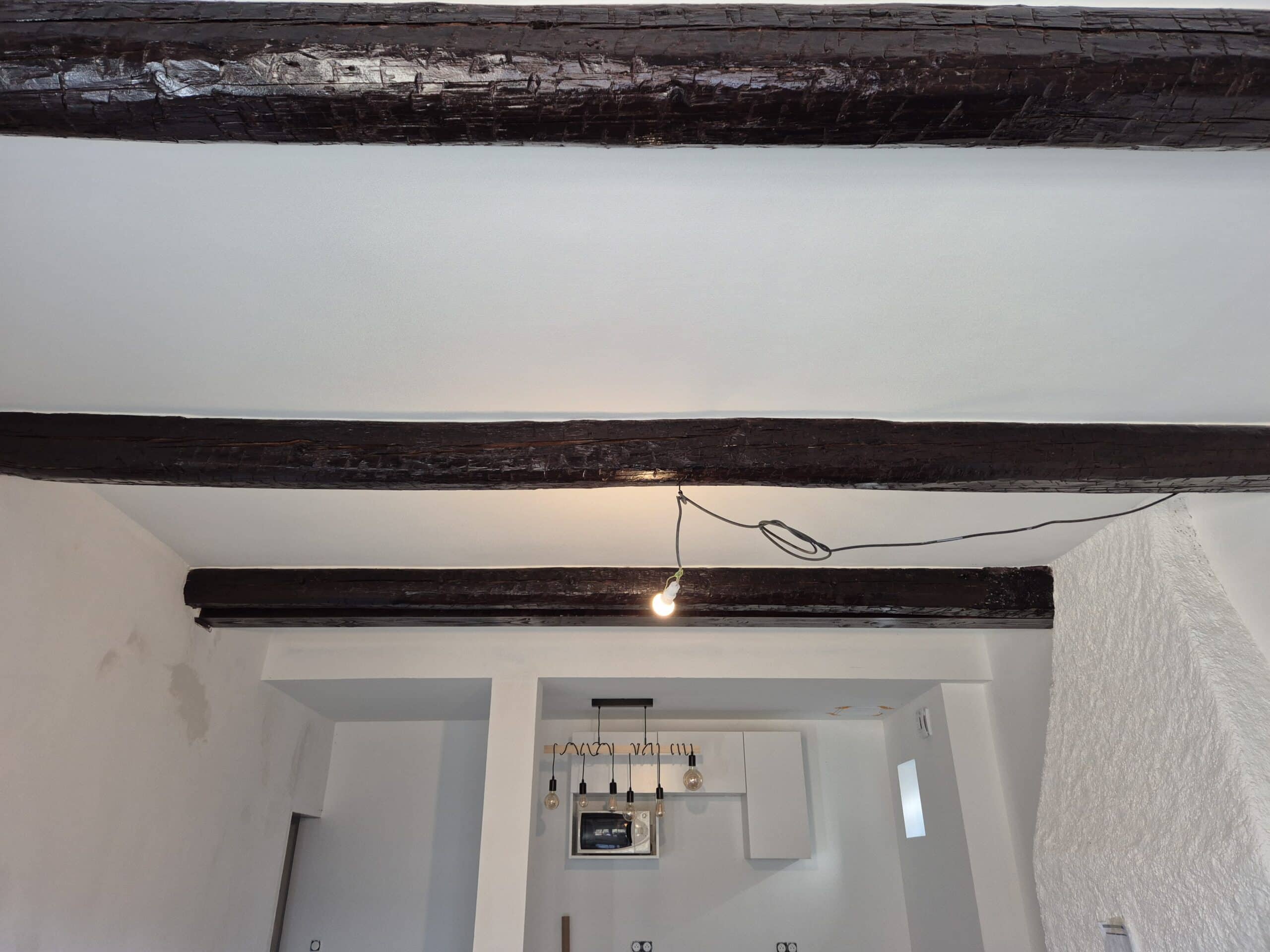 Intérieur en rénovation avec poutres apparentes foncées, plafonds blancs et éclairage temporaire par ampoule nue.