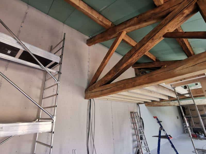 Chantier rénovation grange : murs en plaques de plâtre, charpente apparente en bois massif, échafaudage métallique et échelles visibles.