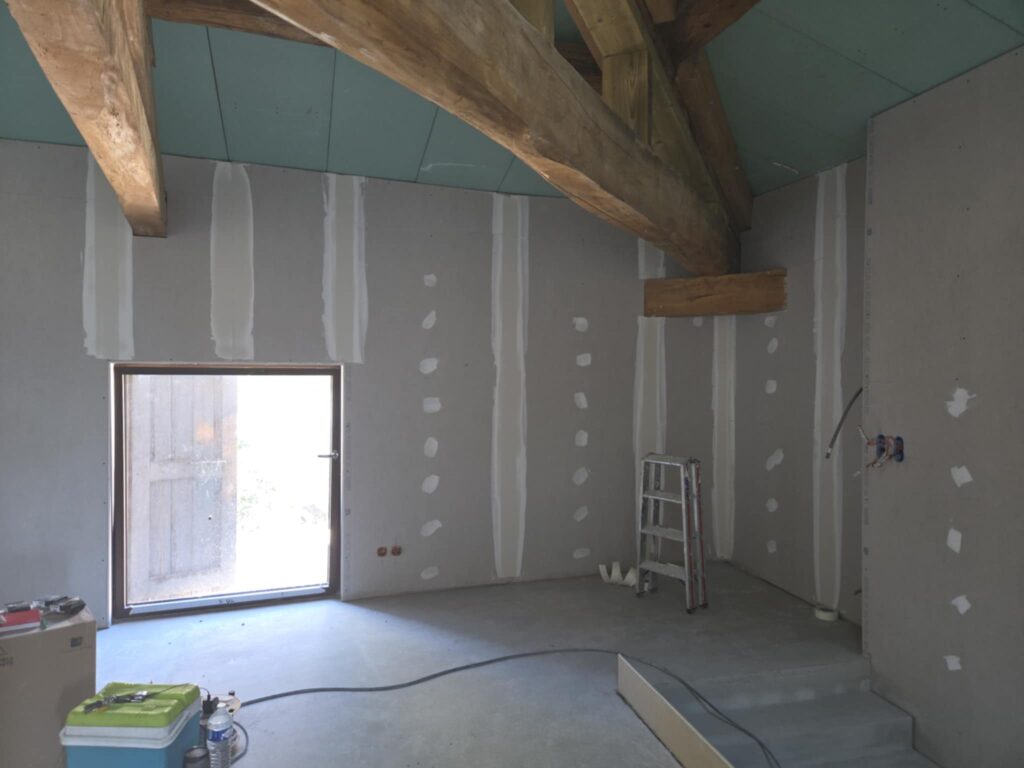 Rénovation Placo et Poutres : Chantier Intérieur en Cours Intérieur d'une pièce en rénovation avec plaques de plâtre jointoyées, poutres apparentes en bois, et une porte-fenêtre lumineuse. Matériel de chantier au sol.