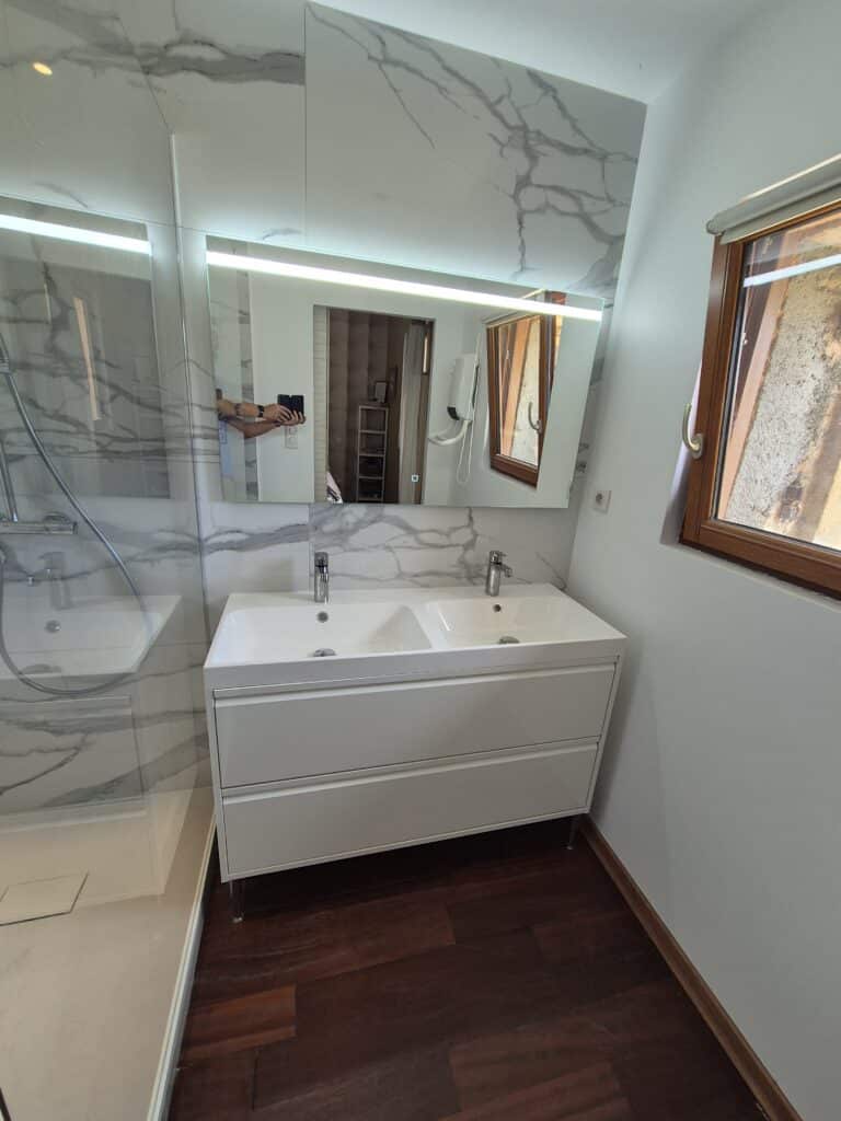 Salle de Bain Design : Double Vasque et Douche Salle de bain moderne avec meuble-lavabo double blanc, miroir éclairé, douche vitrée et sol en bois foncé. La personne prend la photo.