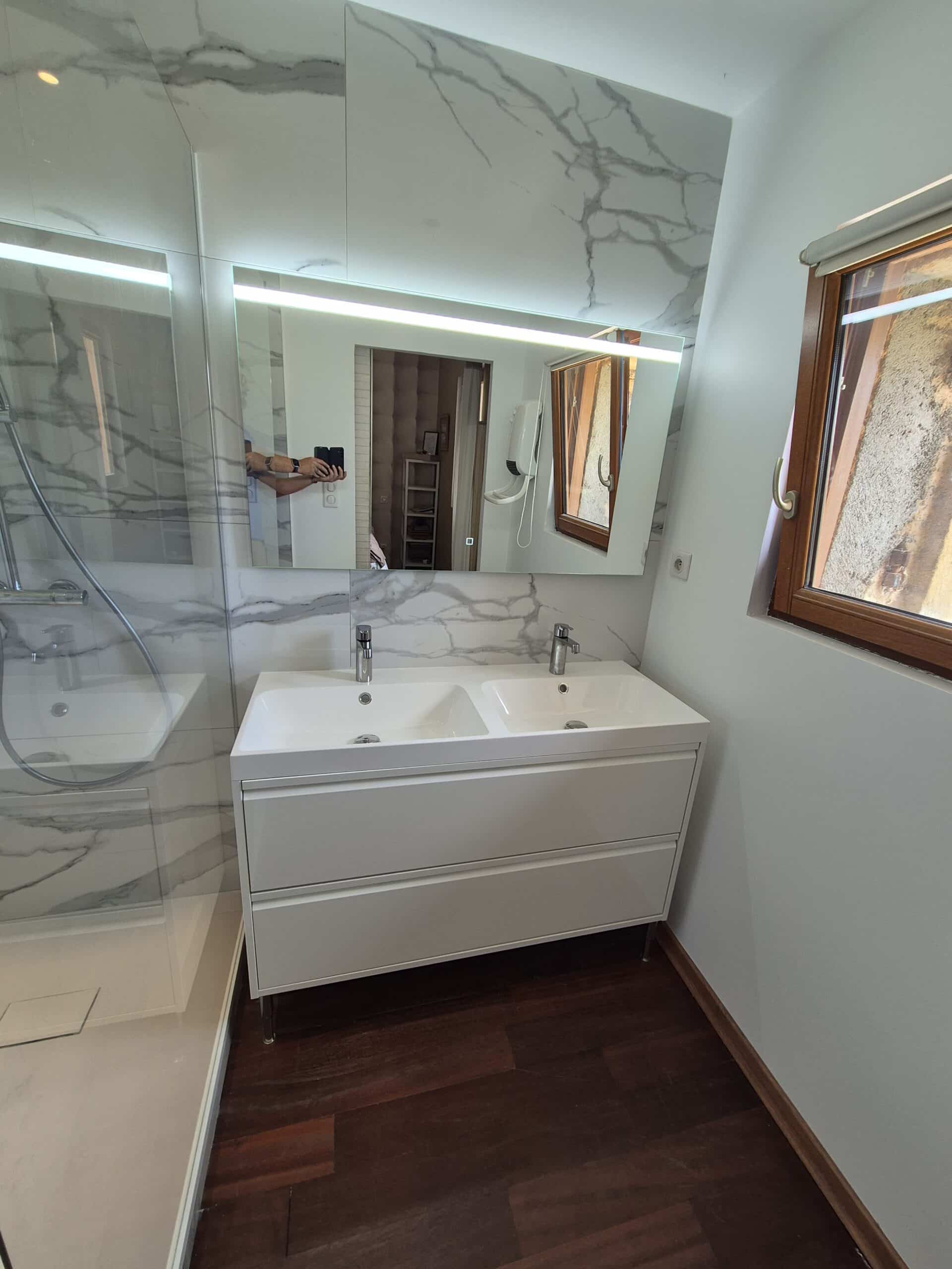 Salle de bain moderne avec meuble-lavabo double blanc, miroir éclairé, douche vitrée et sol en bois foncé. La personne prend la photo.