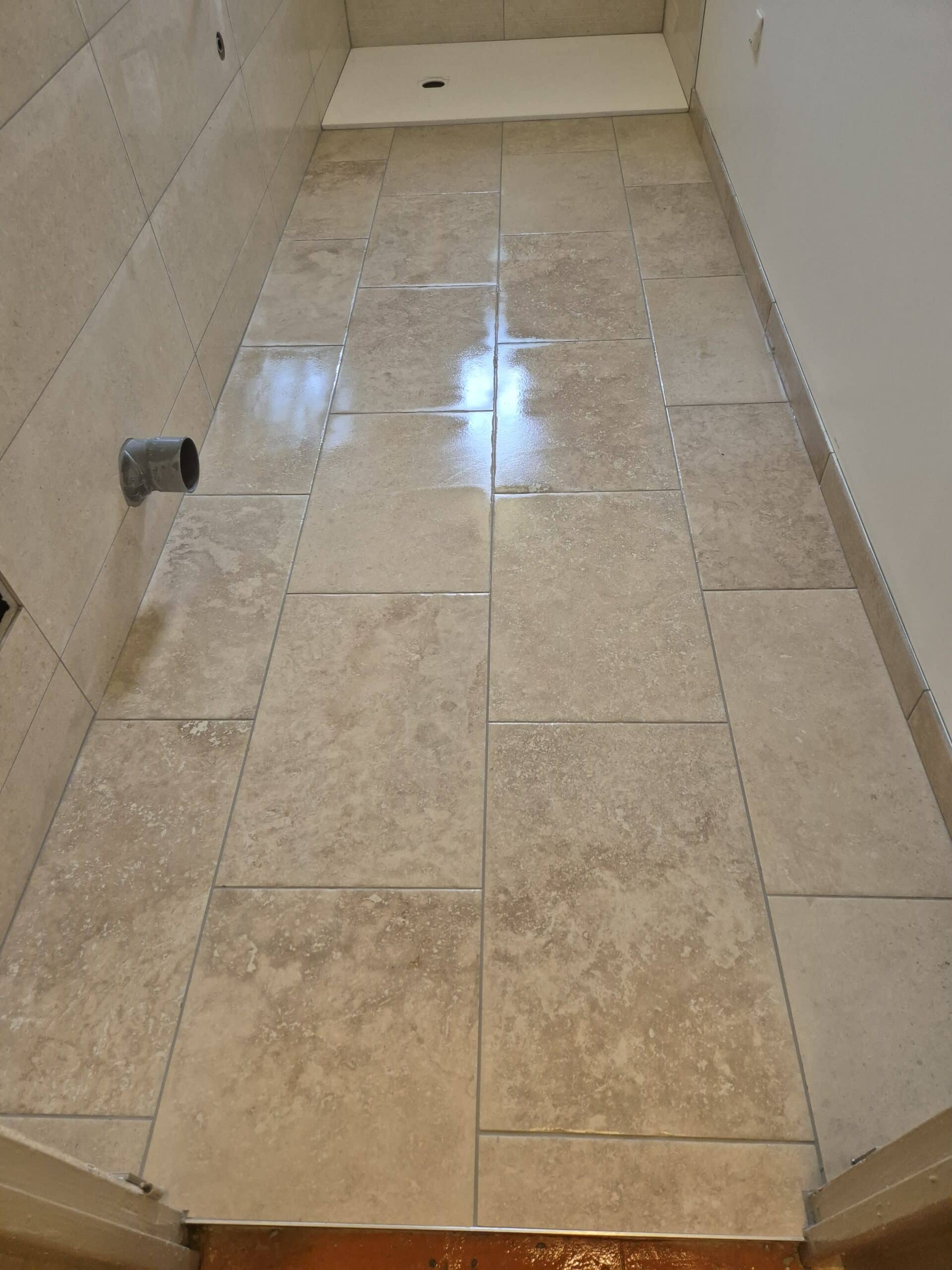 Sol carrelé beige clair de type travertin brillant, avec un drain visible dans le fond et un raccord de tuyau gris sortant du mur.