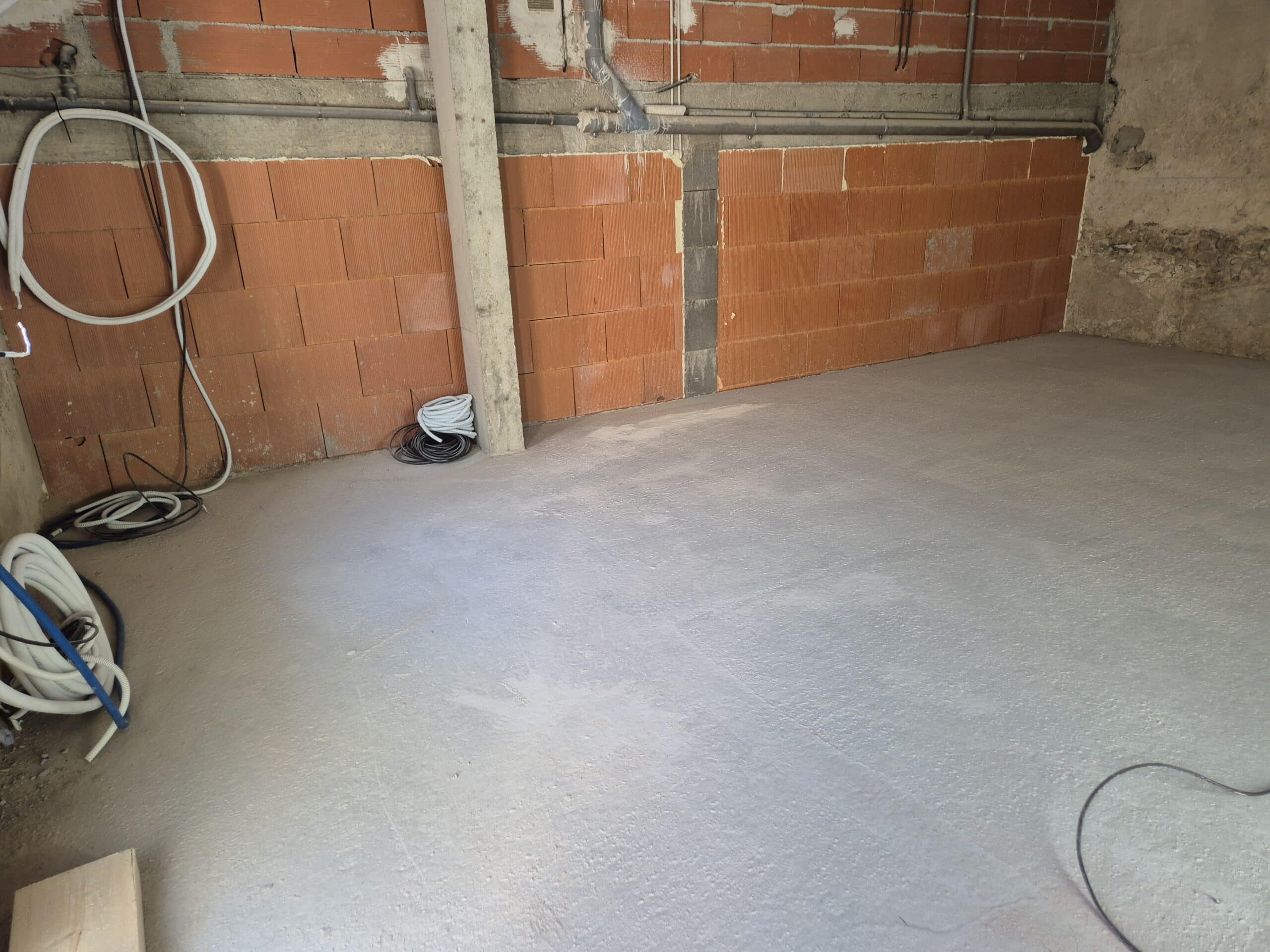 Chantier intérieur avec nouveau sol en ciment gris, murs en briques de terre cuite et tuyauterie/câbles en attente.
